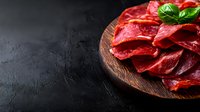Fresh sliced beef carpaccio arranged on wooden plate with basil leaves against dark background, raw meat delicacy for restaurant menu and food service.