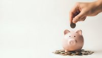 Hand dropping coin into pink piggy bank surrounded by golden coins on white background for savings and financial planning concepts.