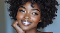 Young African American woman with curly natural hair smiling confidently with glowing skin and perfect makeup against soft blue background.