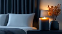 Cozy bedroom interior with burning candle and dried flowers in vase on nightstand. Soft evening light creates relaxing atmosphere against white bedding and dark curtains.