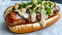Grilled sausage in soft bun topped with creamy sauce, caramelized onions and fresh chopped parsley. Gourmet street food with melted cheese sauce drizzle.