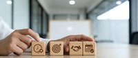 Businessman arranging wooden blocks with business icons including clock, target, handshake, and calendar on office desk, symbolizing strategy and planning.