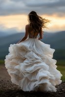 Young woman in flowing white wedding dress with ruffled skirt standing on mountain path at sunset, hair blowing dramatically in wind against stormy sky.