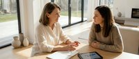 Young adult woman of Caucasian ethnicity in business casual attire discussing documents with female colleague at wooden desk in modern office space with natural light.
