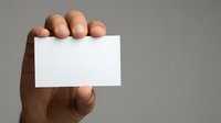 Male hand with medium-dark skin tone holding blank white business card or namecard against neutral gray background for corporate branding and identity mockup purposes.