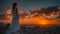 Young woman in flowing white dress standing on rocky cliff overlooking ocean during golden sunset with dramatic orange clouds in sky for travel and lifestyle content.