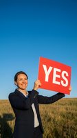 Caucasian male professional in dark business suit holding red sign with white text in open field under clear sky, expressing positive affirmation and agreement for corporate messaging.