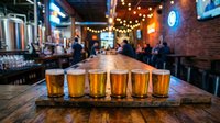 Craft beer flight with multiple golden ales and lagers served on wooden board in busy pub with warm lighting and blurred patrons, perfect for beverage, hospitality and restaurant marketing.
