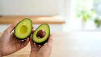 Hands holding halved fresh avocado with pit visible in bright kitchen setting, showcasing ripe green fruit for healthy eating, cooking, nutrition, and food preparation concepts.