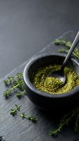 Ground green herbs powder in stone mortar with pestle and fresh thyme sprigs on dark slate surface for cooking and seasoning culinary preparations.