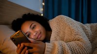 Young woman of mixed ethnicity with curly dark hair, wearing a cozy beige knit sweater, relaxing in bed at night while using smartphone. Warm bedroom lighting.