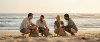 Four diverse young adults sitting cross-legged on sandy beach during golden hour sunset, sharing drinks and conversation by ocean waves in relaxed casual wear.
