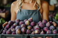 Young Caucasian woman in denim apron holding tray of fresh purple figs at farmers market, showcasing organic seasonal produce for healthy eating.