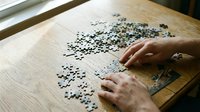 Hands assembling jigsaw puzzle pieces on wooden table surface for leisure activity and mental exercise entertainment.