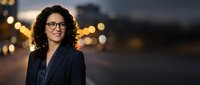 Young woman with curly dark hair and glasses wearing dark blazer against blurred evening cityscape with golden bokeh lights, suitable for corporate, business, and professional portraits.