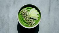 Green smoothie bowl topped with pumpkin seeds and sliced apple, served in white bowl on gray marble surface for healthy breakfast and nutrition content.