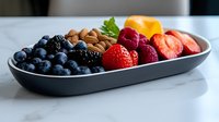 Fresh mixed berries and nuts arranged on modern gray ceramic platter against white marble background, colorful healthy breakfast or snack composition.