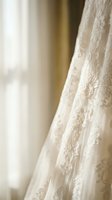 Elegant white wedding dress with intricate lace embroidery and floral patterns hanging in soft natural light for bridal preparation and ceremony planning.