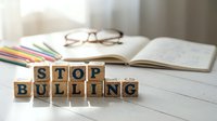 Wooden letter blocks spelling STOP BULLYING on desk with open notebook, glasses and colored pencils in soft natural light for anti-bullying awareness campaigns.