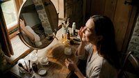 Young woman with long dark hair and light skin tone applying makeup at wooden vanity with round mirror, surrounded by cosmetics and brushes in warm-lit room. Suitable for beauty, skincare, and lifestyle content.
