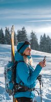 Young blonde woman skier using smartphone on snowy mountain slope with forest background and blue sky for winter sports and technology communication.