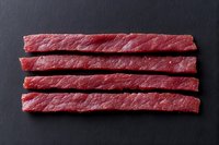 Raw beef strips arranged in parallel rows on dark slate background, fresh meat cuts showing marbling and texture for cooking or restaurant menu presentation.