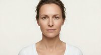 Middle-aged Caucasian woman with natural makeup and brown hair wearing white top against neutral background for skincare and beauty concepts.