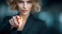 Confident businesswoman in dark suit pointing directly at viewer with determined expression against blurred corporate background.