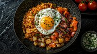 Spicy kimchi fried rice topped with sunny side up egg, crispy bacon, roasted vegetables and fresh herbs served in black bowl on dark stone background.