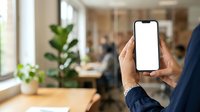 Man in business attire holding smartphone with blank white screen in modern office environment with colleagues and plants visible in background.