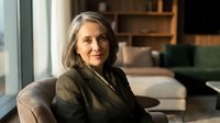 Confident mature businesswoman with gray hair sitting in modern office interior, professional executive portrait for corporate communications.
