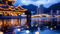 Illuminated traditional Asian pagoda roof with glowing lantern reflections on calm water surface against modern cityscape and mountains at dusk.