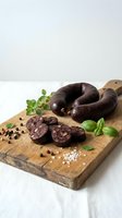 Dark blood sausage slices on rustic wooden cutting board with fresh herbs, sea salt and peppercorns for gourmet cooking and traditional cuisine preparation.