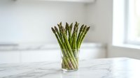 Fresh green asparagus spears standing upright in clear glass vase on white marble countertop in bright modern kitchen interior for healthy cooking.
