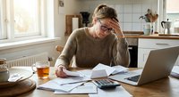 Stressed middle-aged Caucasian woman working from home kitchen table with laptop, papers and tea cup, dealing with financial documents and bills.