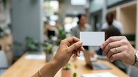 Two professionals exchanging blank white business cards in modern office environment with colleagues and plants in background, networking and corporate communication concept.