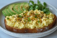 Creamy scrambled eggs on toasted sourdough bread garnished with black sesame seeds, fresh herbs and sliced avocado served on white ceramic plate.