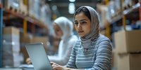 Young Middle Eastern woman in hijab working on laptop in warehouse logistics center, with storage shelves and cardboard boxes in background.