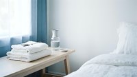 Stacked white towels on wooden nightstand beside bed with water glass and candle, minimalist bedroom interior design.