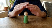Protective hands forming shelter over small wooden peg person figurine on table symbolizing care, security, insurance and human protection concepts.