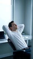 Young Caucasian businessman relaxing in office chair, hands behind head, gazing upward thoughtfully near bright window with laptop on desk.