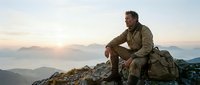 Mature man with dark hair and beard, wearing olive-green hiking shirt and backpack, sitting on rocky mountain summit at sunrise with misty valleys below. Adventure and outdoor lifestyle.