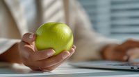 Woman holding fresh green apple while working on laptop promoting healthy eating habits and wellness lifestyle in modern office environment.
