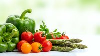 Fresh organic vegetables including green bell peppers, cherry tomatoes, and asparagus spears arranged on white surface with natural lighting for healthy cooking.