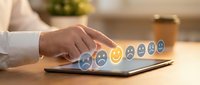 Person with medium skin tone selecting yellow smiley face on laptop keyboard displaying emotion feedback icons, representing customer satisfaction survey and service rating evaluation.