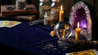 Mystical fortune telling setup with crystal ball, amethyst geode, burning candles, tarot cards and ancient books on dark blue velvet cloth for divination.