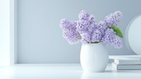 Fresh lilac flowers in white ceramic vase against light wall with stacked books, minimalist spring home decor composition in soft pastel colors.