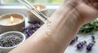 Hand applying clear serum from glass dropper on skin with lavender flowers and candle creating spa wellness skincare routine atmosphere for natural beauty treatment.