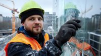 Caucasian male construction worker in safety vest and hardhat cleaning glass facade of modern building, urban cityscape with cranes in background.