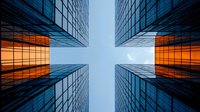 Modern glass skyscrapers photographed from below against blue sky, with dramatic orange sunset reflection in windows creating cross-shaped negative space.